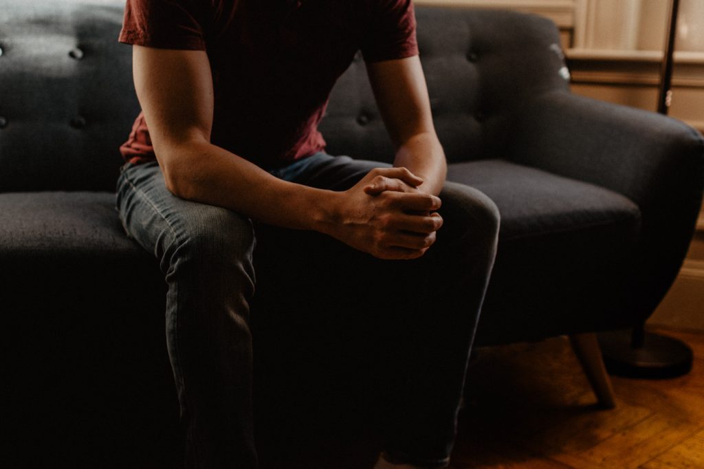 A worried person sitting on a sofa, holding their own hands.