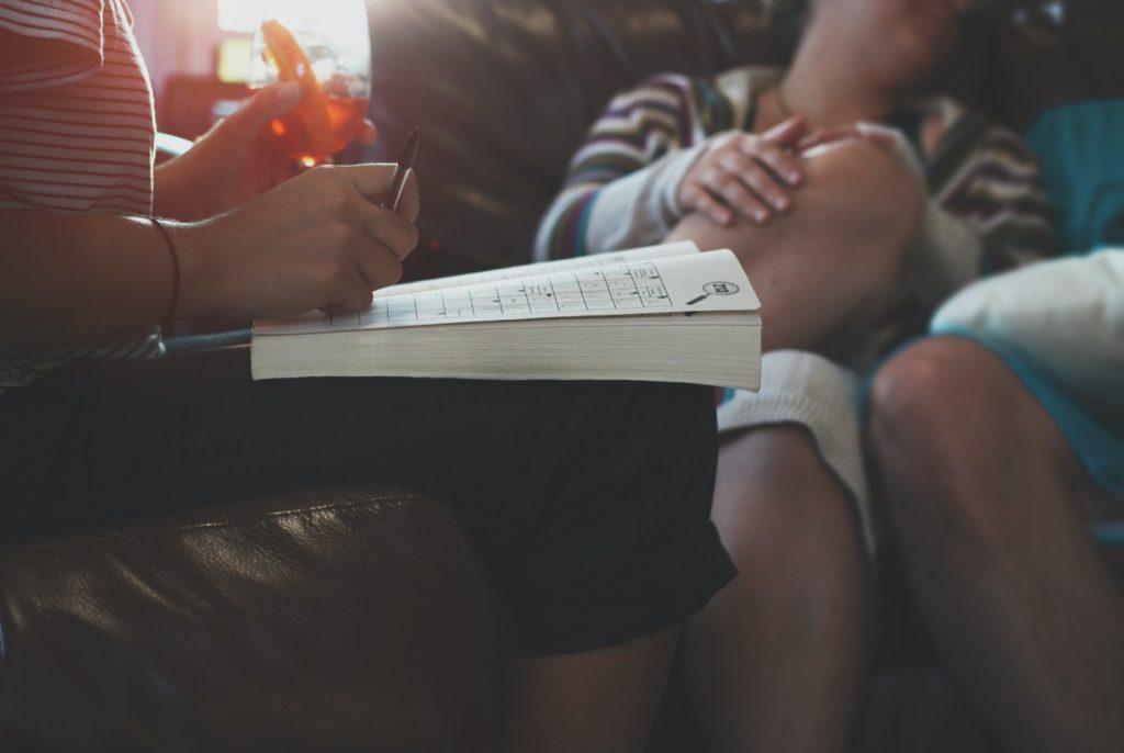 A person writing in a notebook next to two people laughing and enjoying on a sofa.