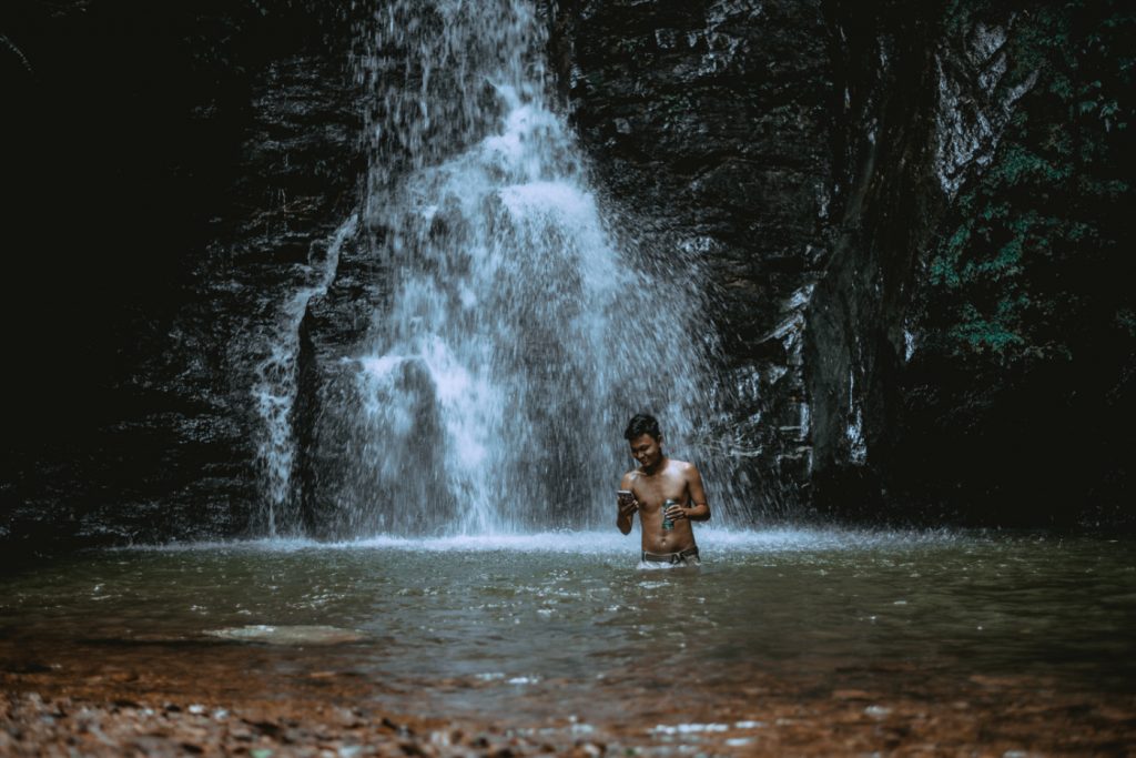 A person bathing next to a waterfall holding a mobile phone and a beer.