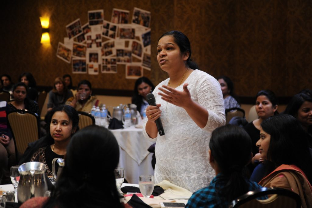 A woman standing up holding a microphone and asking a question while people beside her watch and listen.