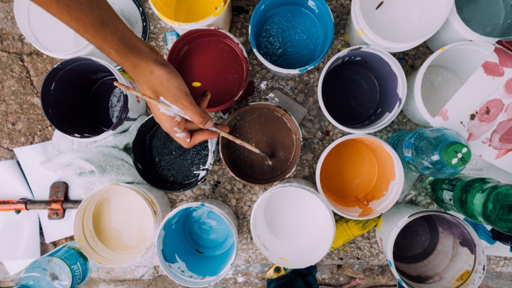 A person dipping a brush into a paint pot among many different colors.