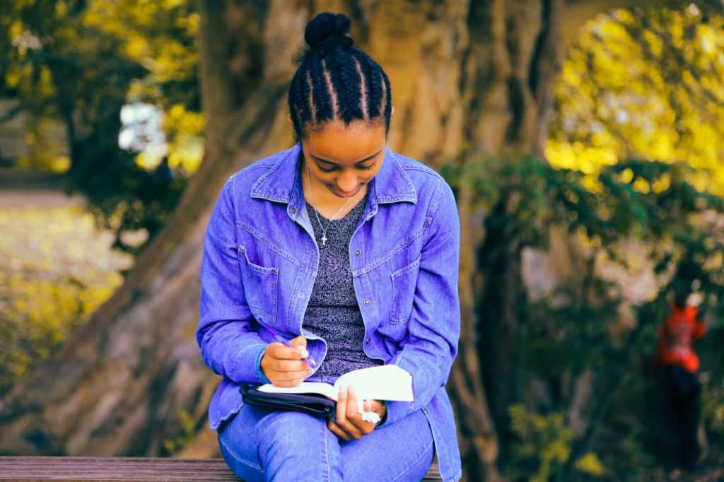 A woman sitting on a bench in a park writing in a notebook.