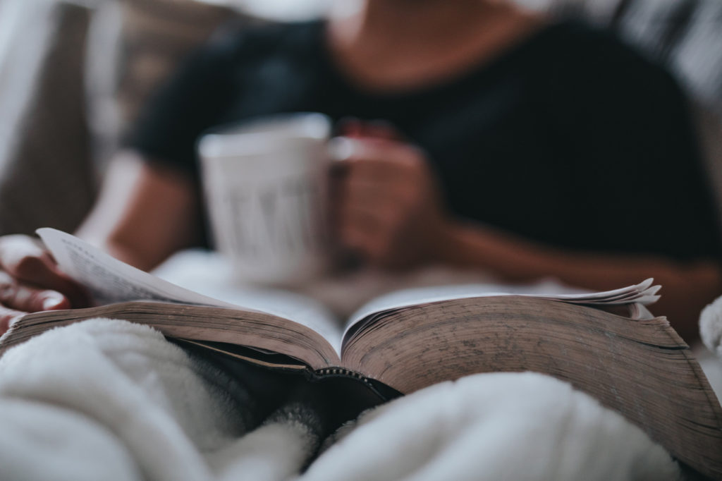 A person holding a cup of tea, reading a book on a sofa covered with a blanket