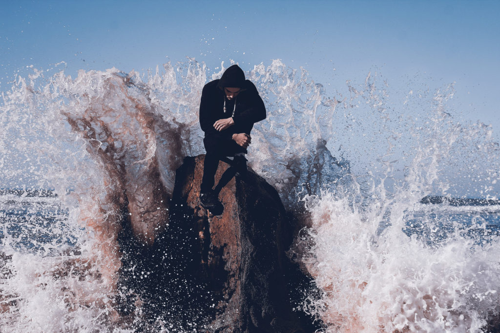 A large wave hitting a man sitting on a rock formation at sea.