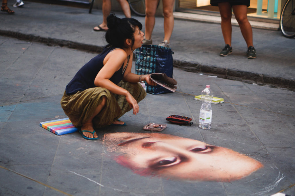 A person creating artwork on the pavement among a couple of bystanders.