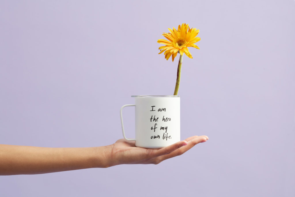 A person holding a mug with a yellow flower peaking out of it and transcription: "I am the hero of my own life."